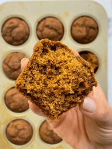 Someone's hand holding a broken open muffin with a pan of muffins in the background. 